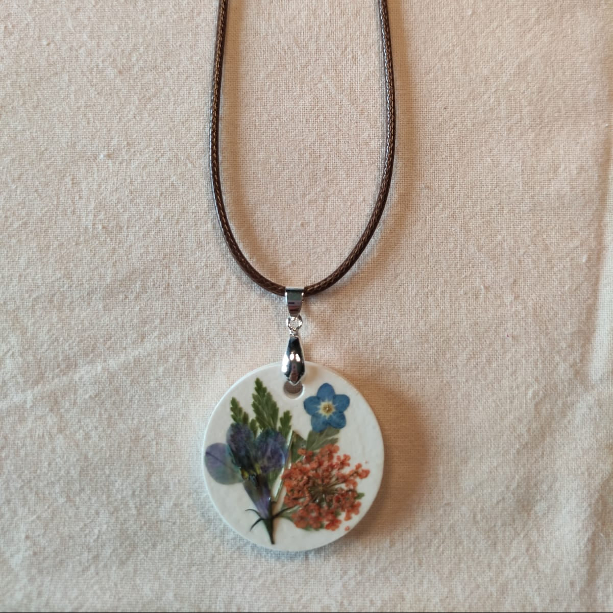 Collier médaillon rond en pierre blanche orné de myosotis bleu, d'une ombelle orangée et de fougère séchée, monté sur cordon ciré brun — NERÉA