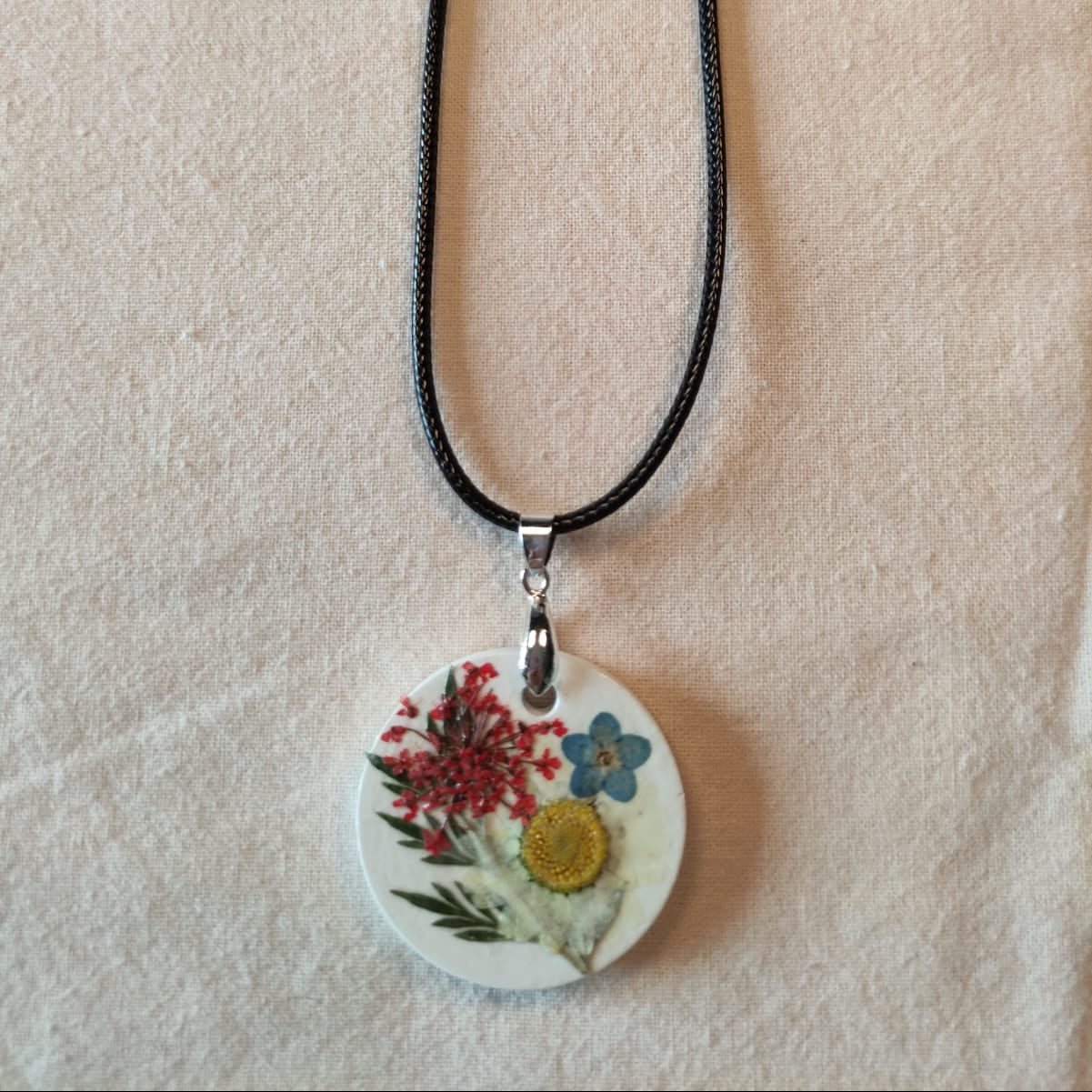 Collier médaillon rond en pierre blanche orné d'une ombelle rouge, d'un myosotis bleu et d'une fleur blanche à cœur jaune séchés, monté sur cordon ciré noir — NERÉA
