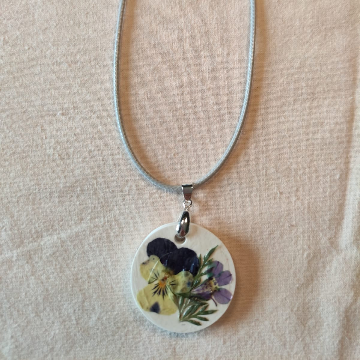 Collier médaillon rond en pierre blanche orné d'une pensée jaune pâle à pétales violets et de fleurs mauves séchées, monté sur cordon ciré gris perle — NERÉA